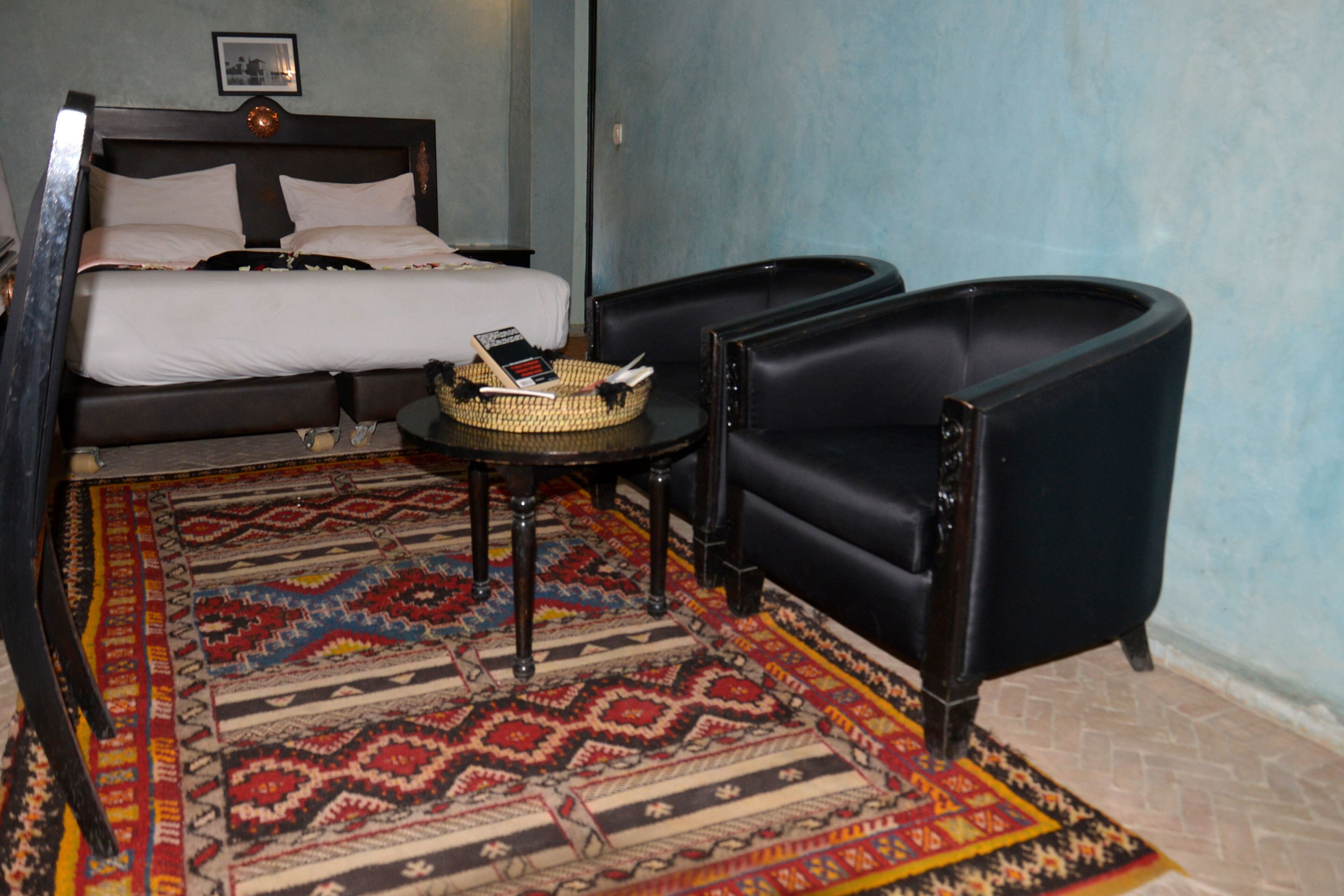 Une pièce comprenant un lit, deux fauteuils noirs, une petite table ronde surmontée d’un panier en osier et un tapis à motifs.