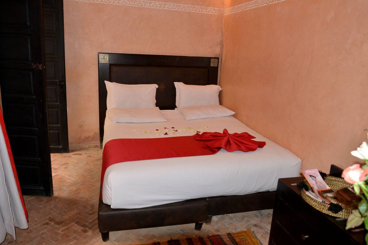 A neatly made bed with white linens and a red decorative bow, a dark wooden headboard, and a small bedside table with a basket.