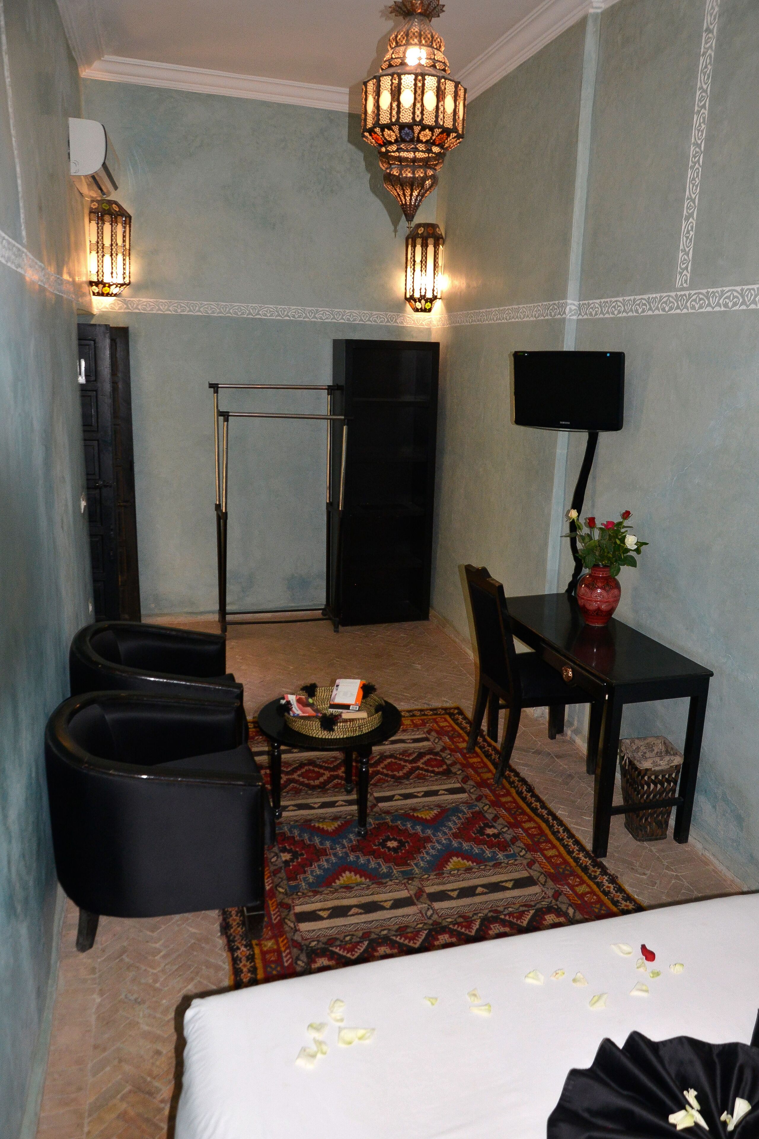 A hotel room with a bed, a small table with a vase of flowers, a chair, a sofa, a television, and a patterned rug.