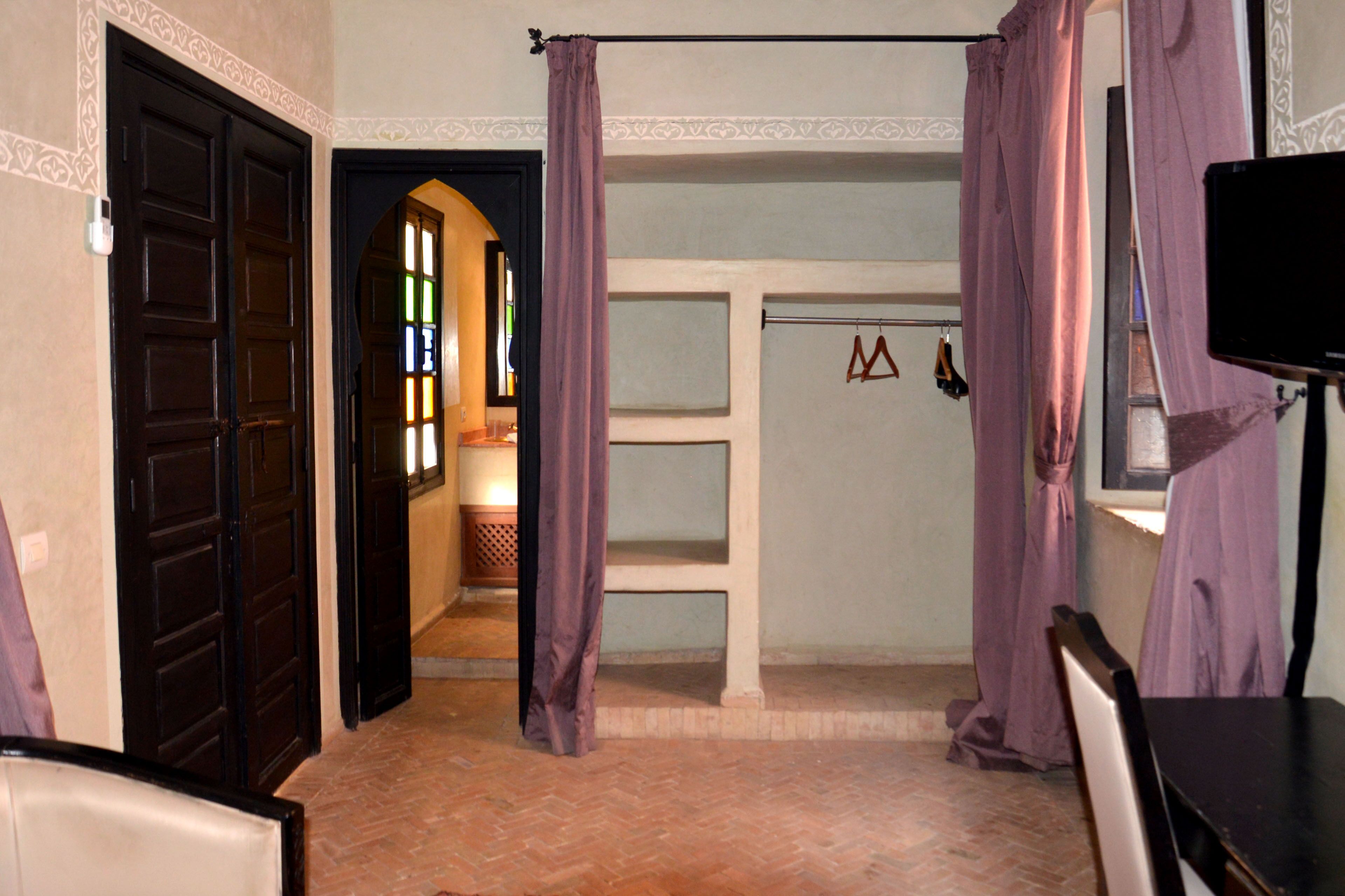 Una habitación con piso de ladrillo, un televisor negro sobre un pedestal y una puerta que da a un pasillo con ventana.
