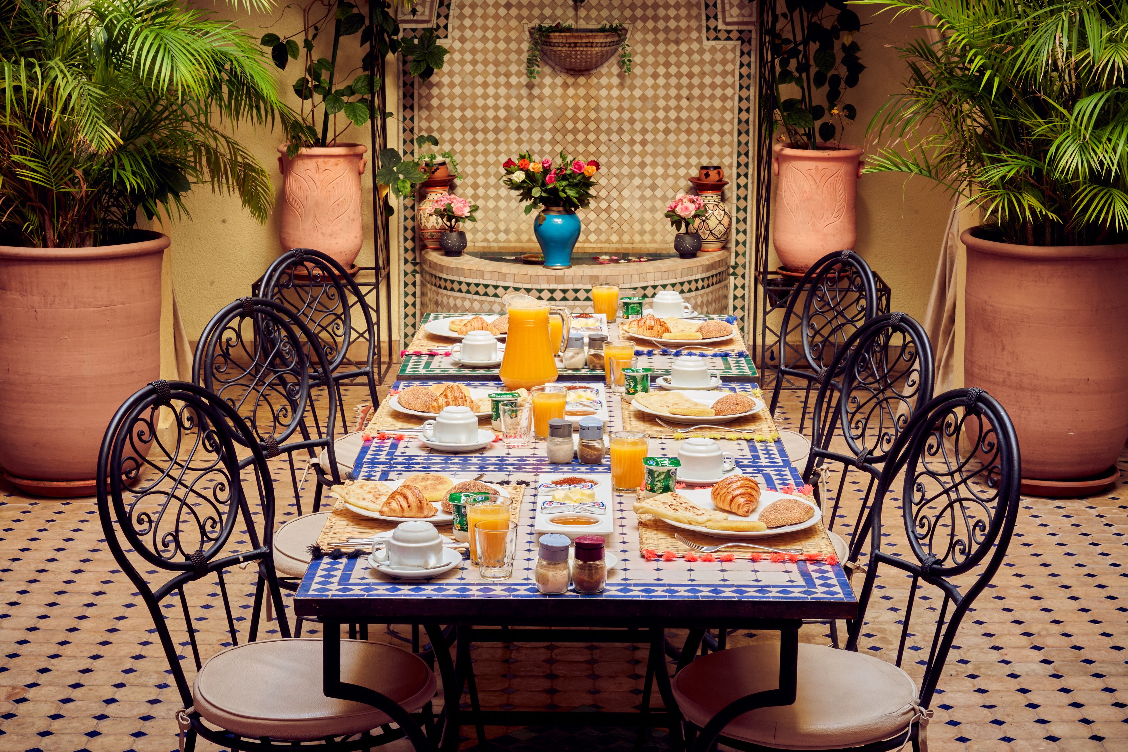 Une table à manger dressée avec des aliments pour le petit-déjeuner, entourée de chaises, dans une pièce dont le sol est recouvert de carreaux décoratifs et où l’on trouve des plantes en pot.