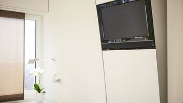 Baño con lavamanos blanco, espejo, orquídea en maceta, toallero y televisor de pantalla plana montado en la pared.