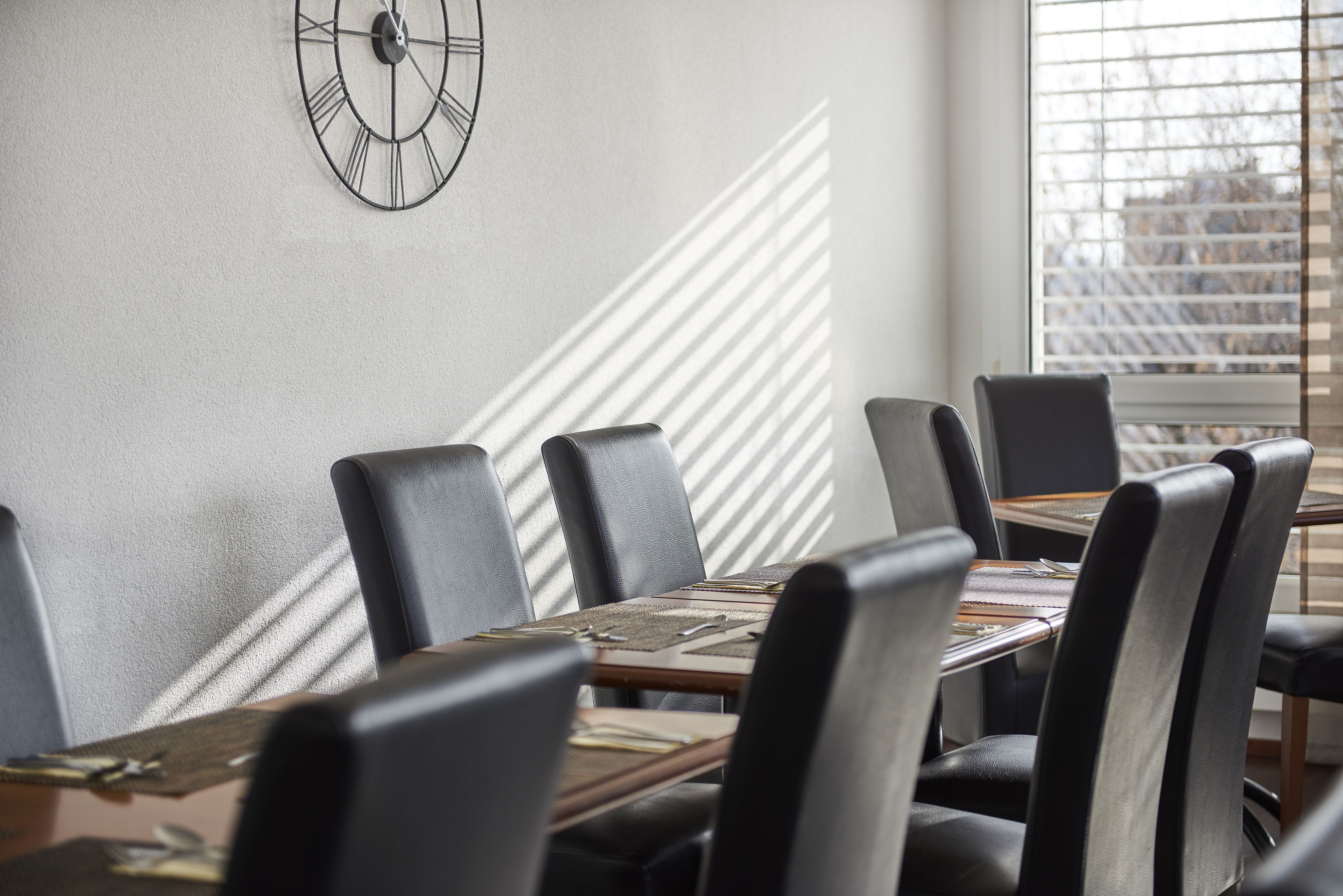 Une salle de conférence avec une horloge au mur, une longue table entourée de chaises et une fenêtre équipée de stores.