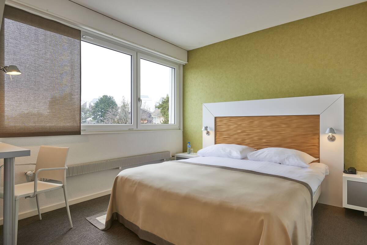 Ein Hotelzimmer mit einem großen Bett, einem Sessel, einem Schreibtisch und einem Fenster mit Blick auf Bäume und Gebäude.