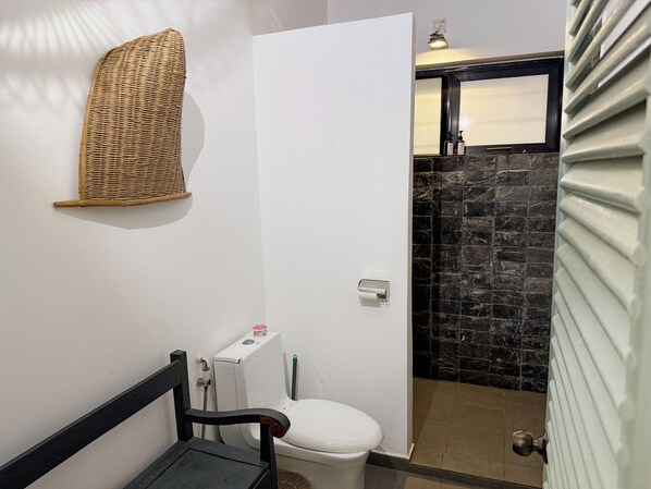 Un baño con un inodoro blanco, un estante de mimbre, una silla negra y una pared de piedra.