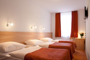 A hotel room with three beds, a wooden headboard, a desk with a chair, a TV mounted on the wall, and a window with orange curtains. - Hotel Ambiance (Prague)