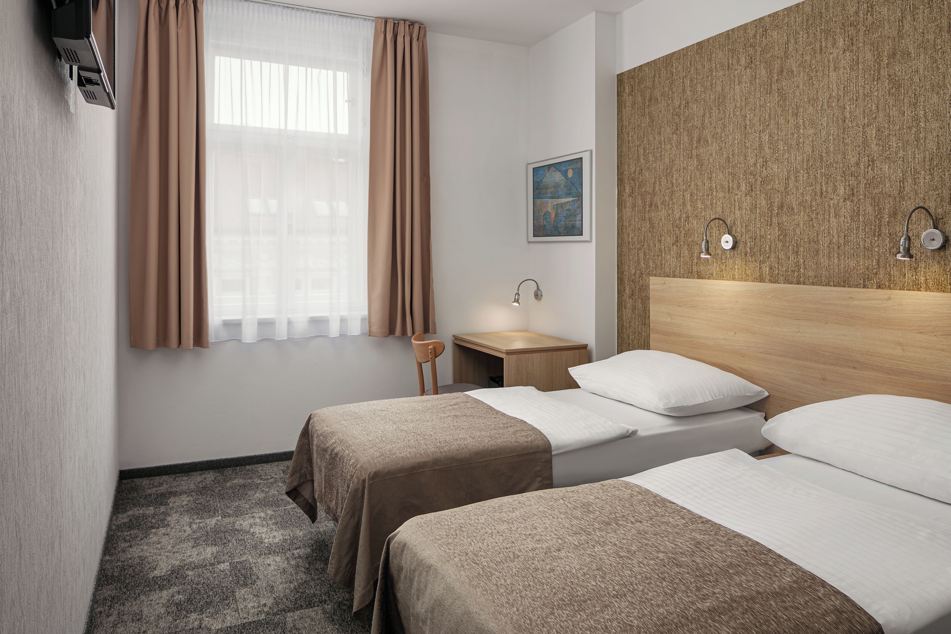 Hotelkamer met twee bedden, een houten hoofdbord, een klein nachtkastje, een stoel, een raam met gordijnen, een schilderij aan de muur en een tv aan de wand.
