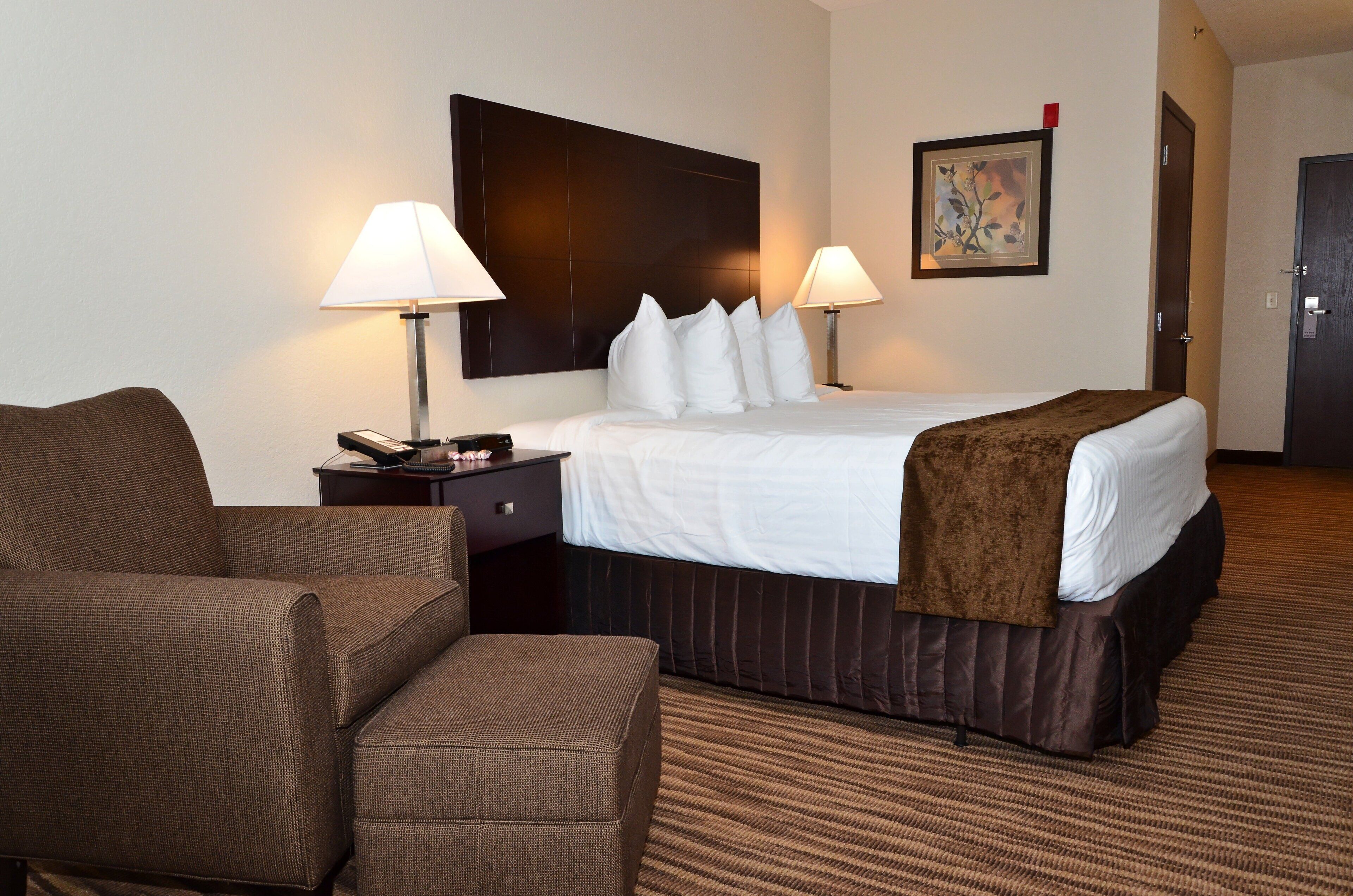 A hotel room with a large bed, two bedside lamps, a brown armchair, a small table with a phone and remote control, and a painting on the wall.
