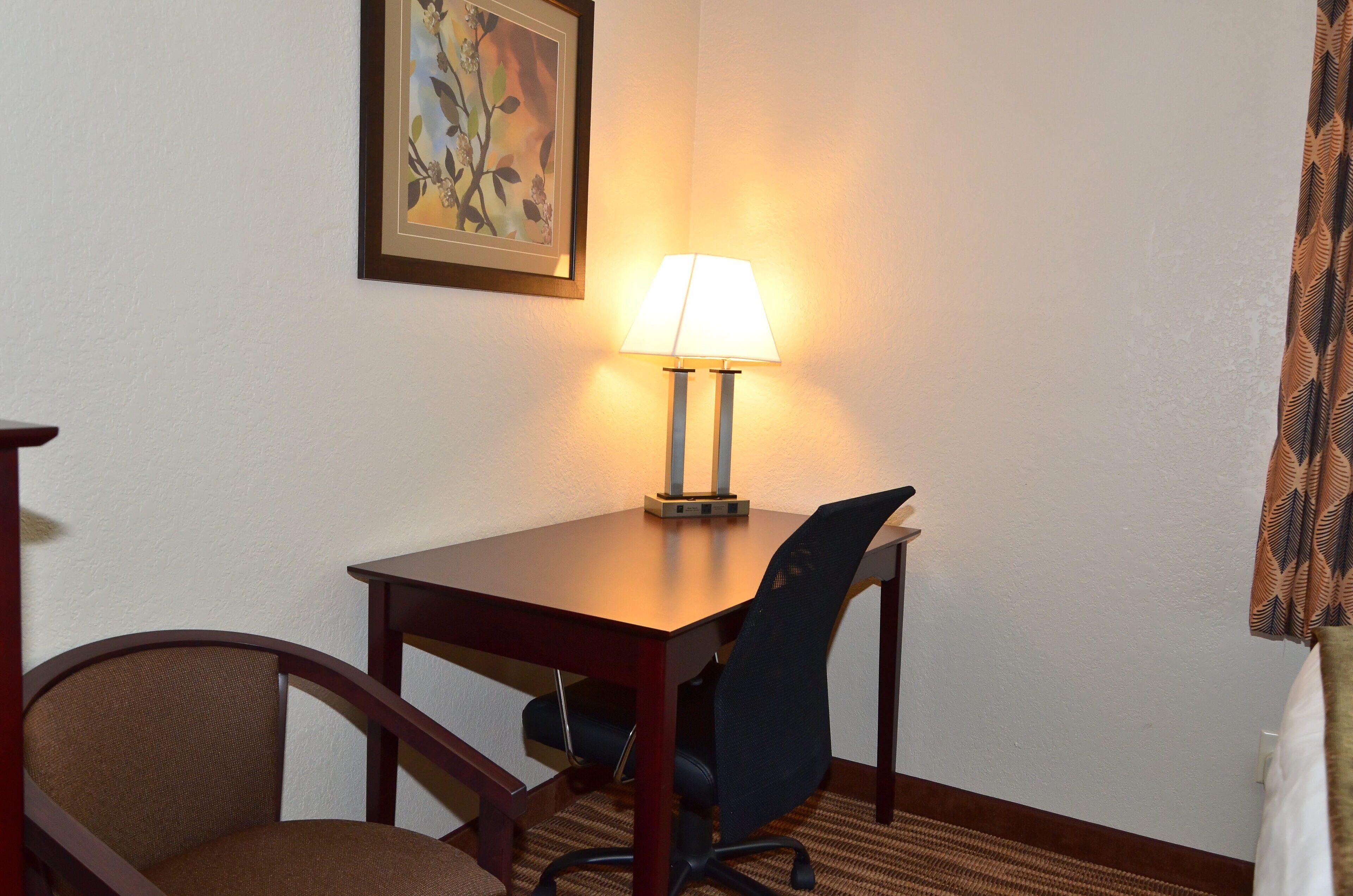 A hotel room with a wooden table, a single chair, a lamp, and a framed picture on the wall.