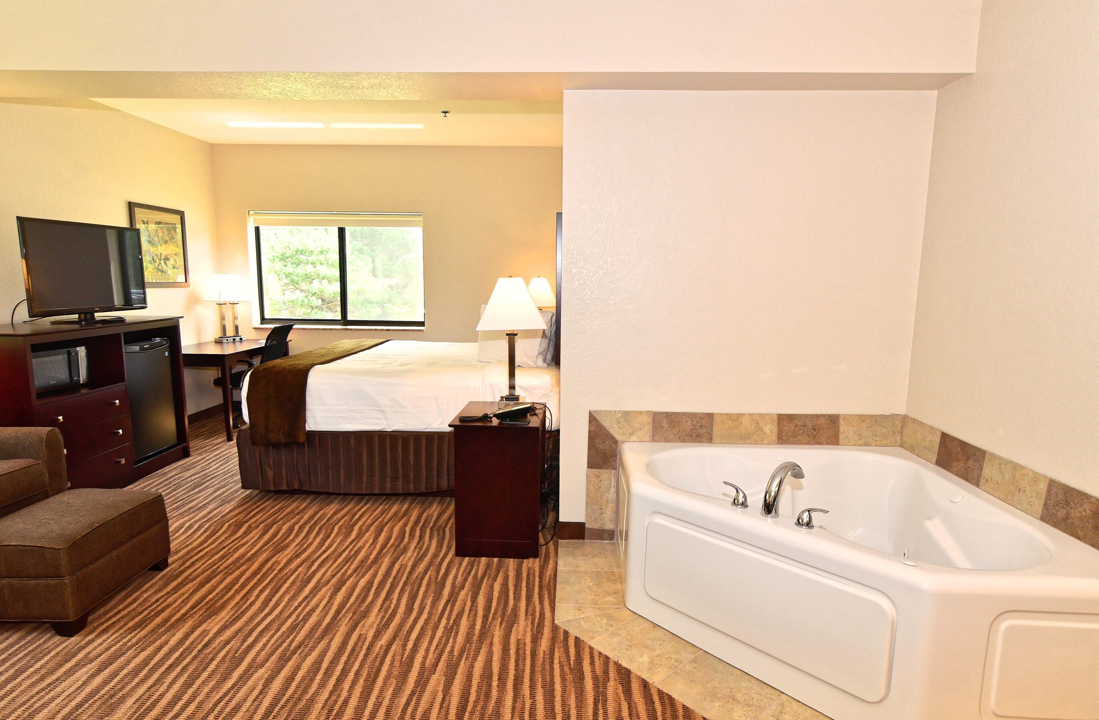 A hotel room with a bed, a desk with a computer, a TV, a sofa, a carpeted floor, and a bathtub.