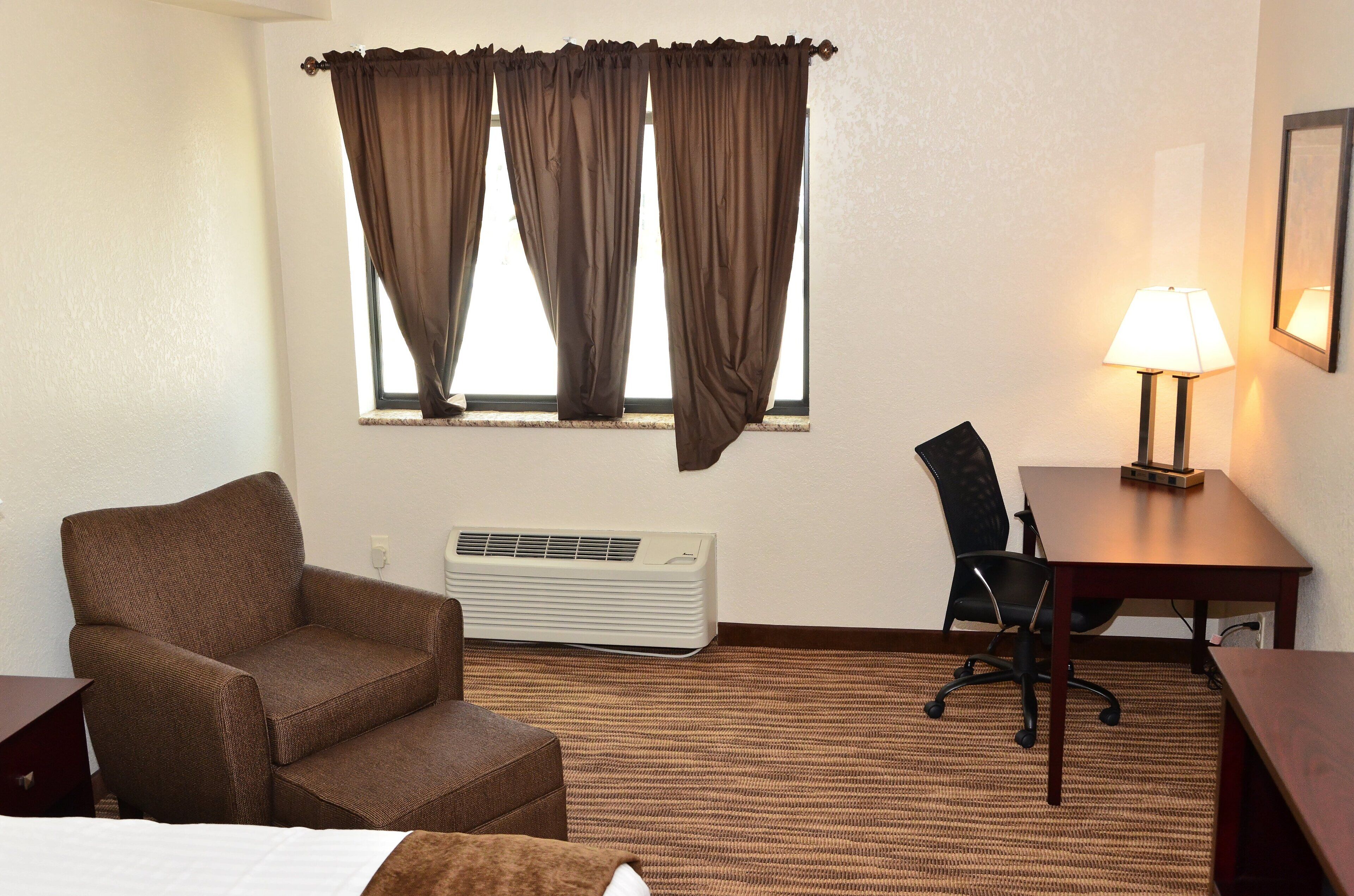 A hotel room with a brown armchair, a desk with a chair, a lamp, and an air conditioning unit.