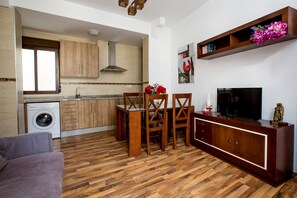 A modern kitchen with wooden cabinets, a built-in oven, and a washing machine. There is a dining area with a table and chairs, and a television on a wooden cabinet.