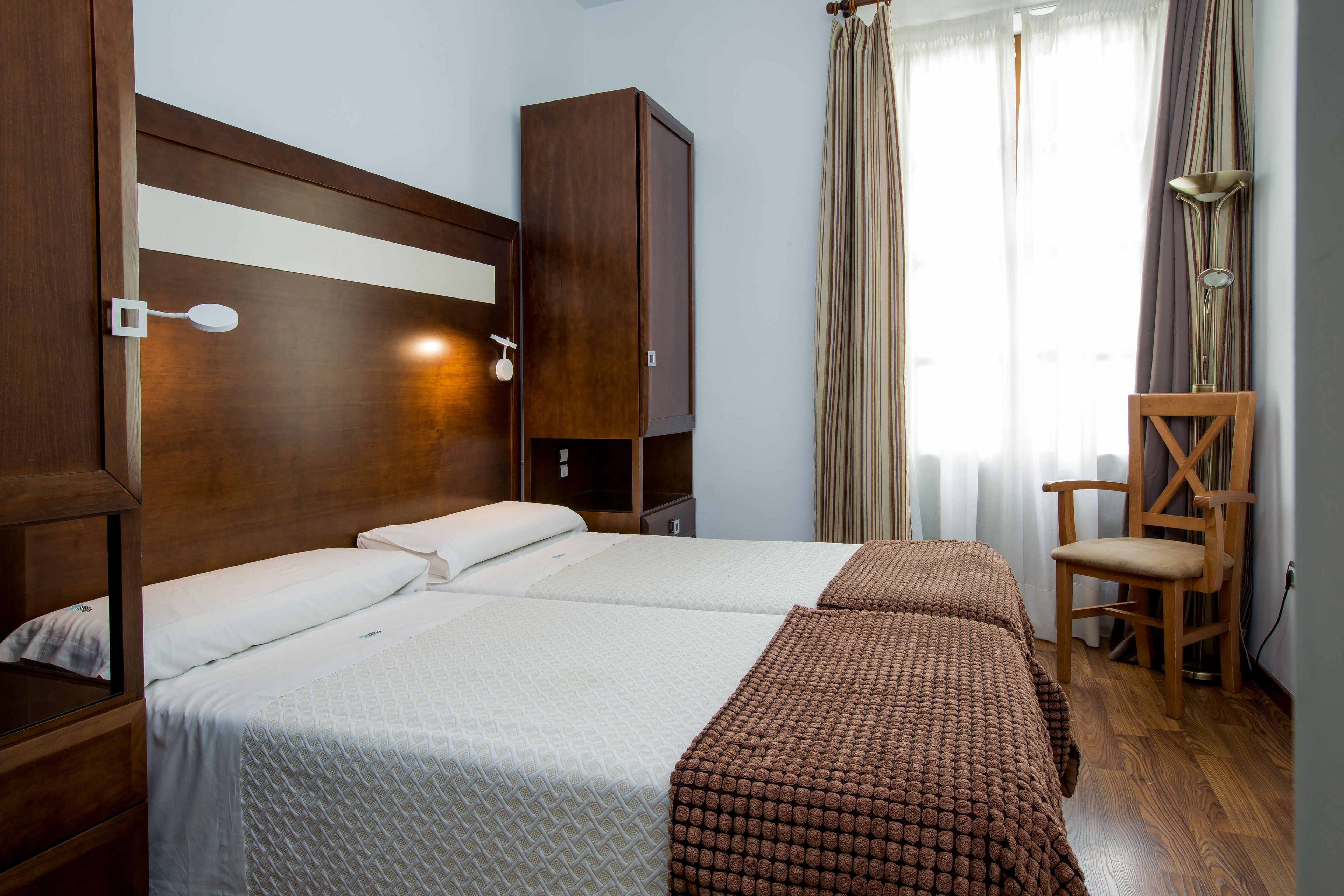 Ein Hotelzimmer mit hölzernem Kopfteil, zwei Betten mit weißer und brauner Bettwäsche, einem Holzschrank und einem Stuhl.