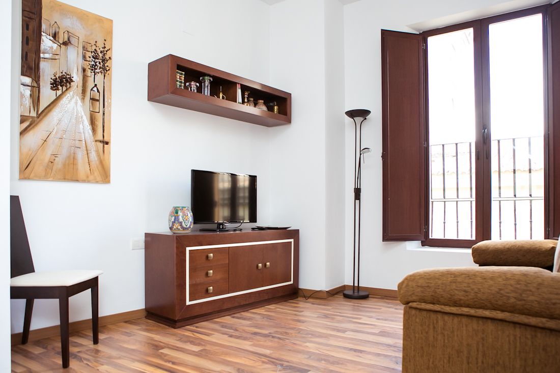 A living room with a wooden floor, a TV on a cabinet, a painting on the wall, and a window with a view.