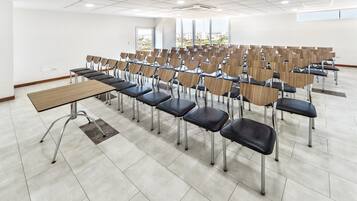 Un salón de conferencias con filas de sillas y una larga mesa, ventanas amplias con vistas a la ciudad, y suelo de baldosas.