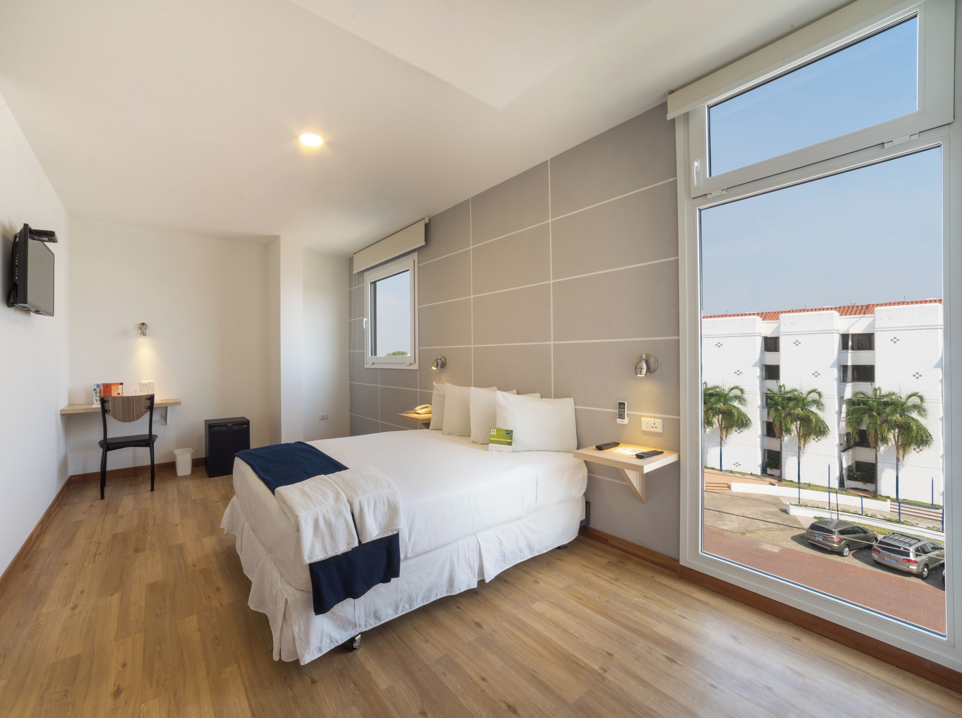 Una habitación de hotel moderna con una cama grande, un escritorio, una silla, un televisor y vistas a palmeras y un edificio.