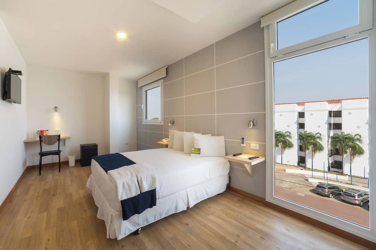Une chambre d’hôtel moderne dotée d’un grand lit, d’un bureau, d’une chaise, d’une télévision et offrant une vue sur des palmiers et un immeuble.