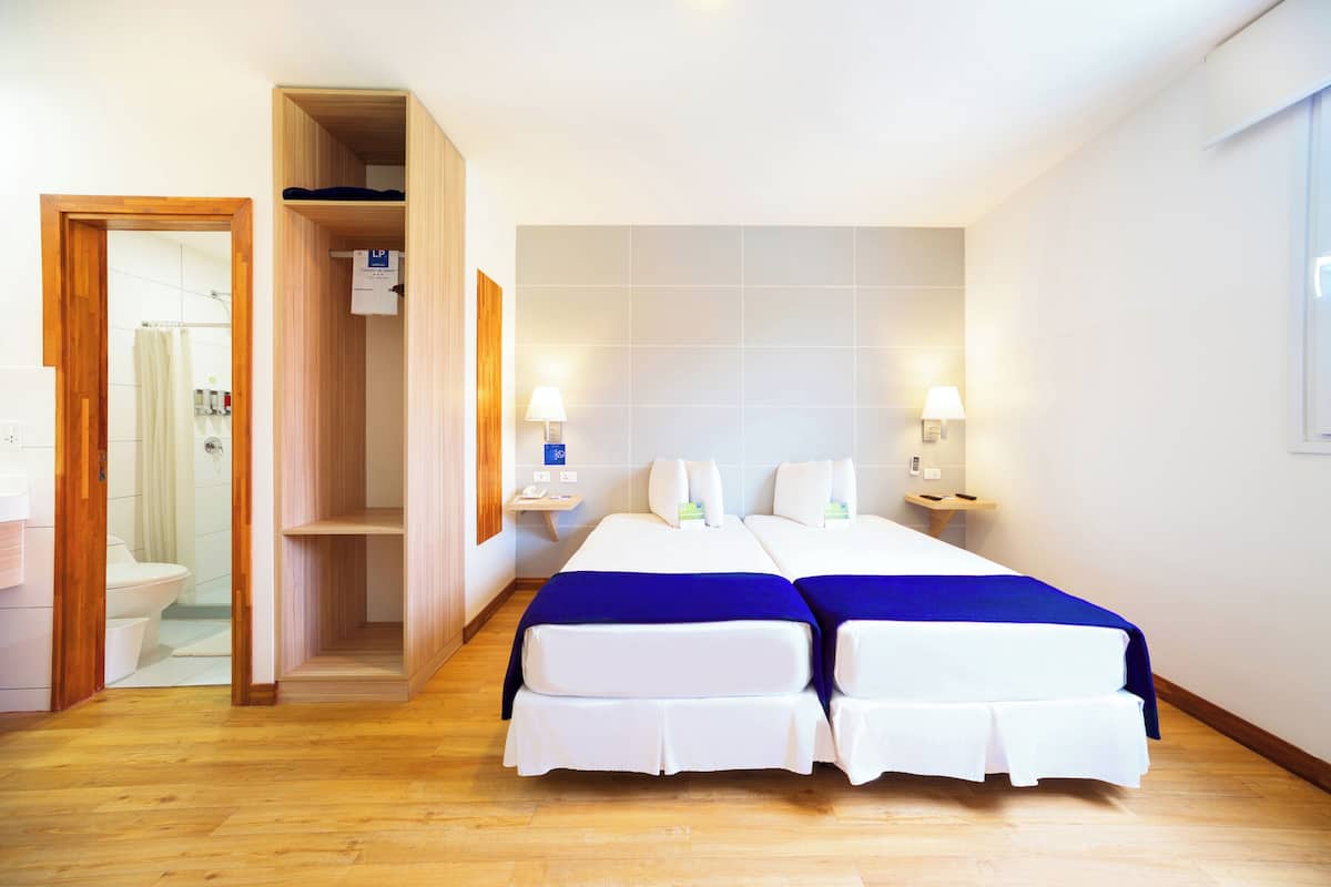 Une chambre d’hôtel avec deux lits, un plancher en bois, une salle de bain avec des toilettes et une douche, et deux lampes de chevet.
