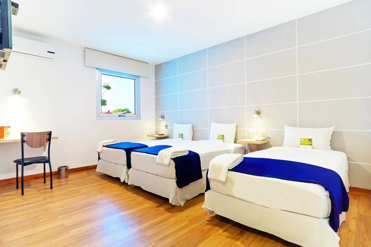 A hotel room with two beds, a desk, a chair, and a window with a view of greenery.
