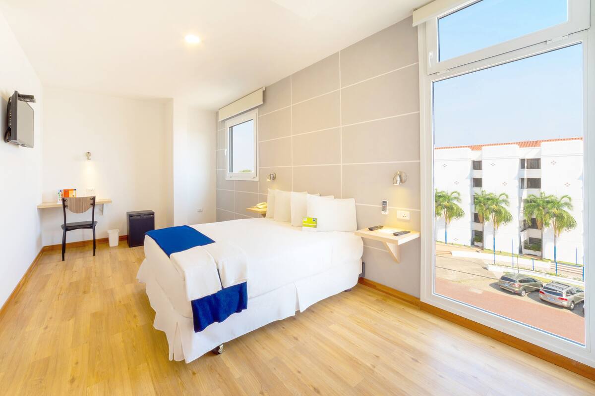 A modern hotel room with a large bed, a desk, a chair, and a view of the outside.