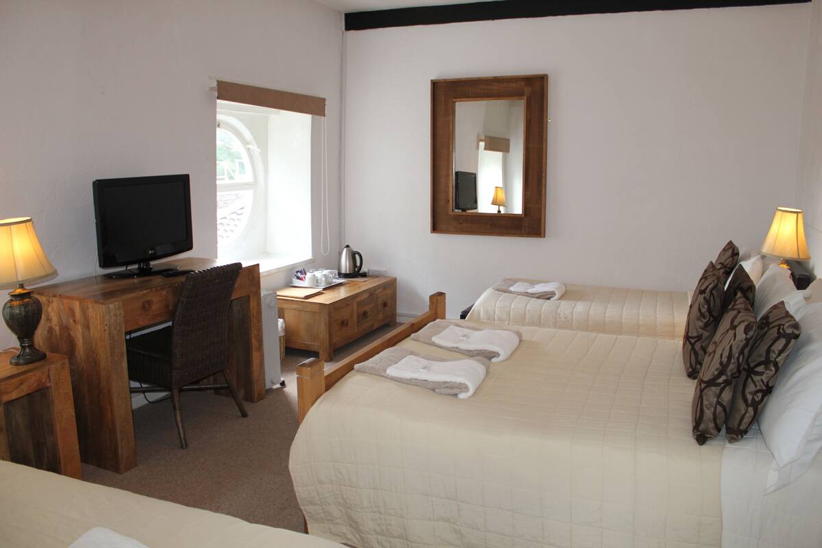 Quarto de hotel com cama, escrivaninha com computador, cadeira, cômoda de madeira, espelho e duas luminárias.