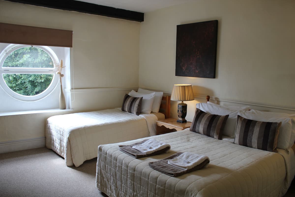 Quarto de hotel com duas camas, janela com vista para área verde, abajur na mesa de cabeceira e um quadro na parede.