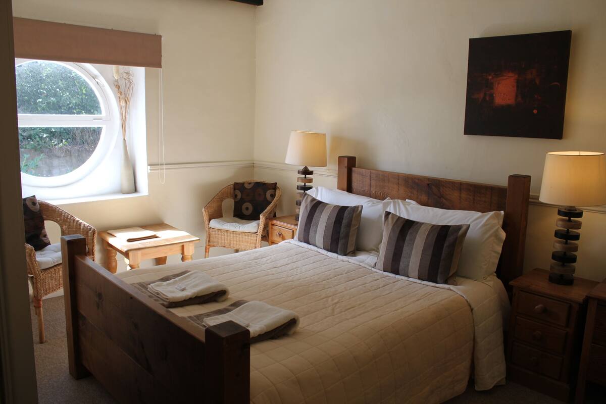 Quarto bem arrumado com uma cama grande, duas mesas de cabeceira com luminárias, uma cadeira e uma janela com vista para o verde.