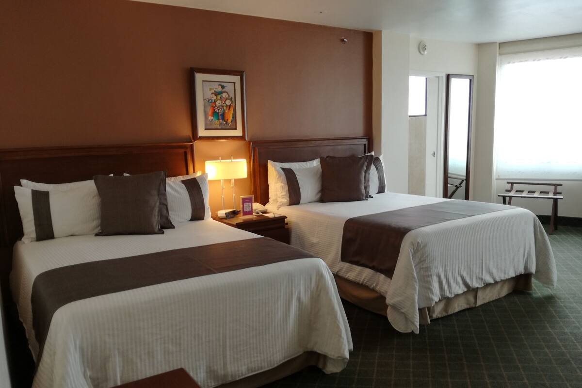 Habitación de hotel con dos camas, una mesita de noche con lámpara, un cuadro en la pared y una ventana con vistas.