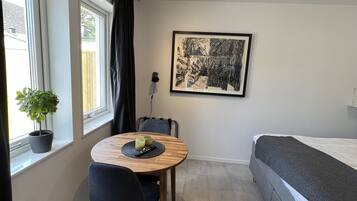 A compact room with a bed, a dining table with chairs, a potted plant, a framed artwork, and a ceiling light fixture.