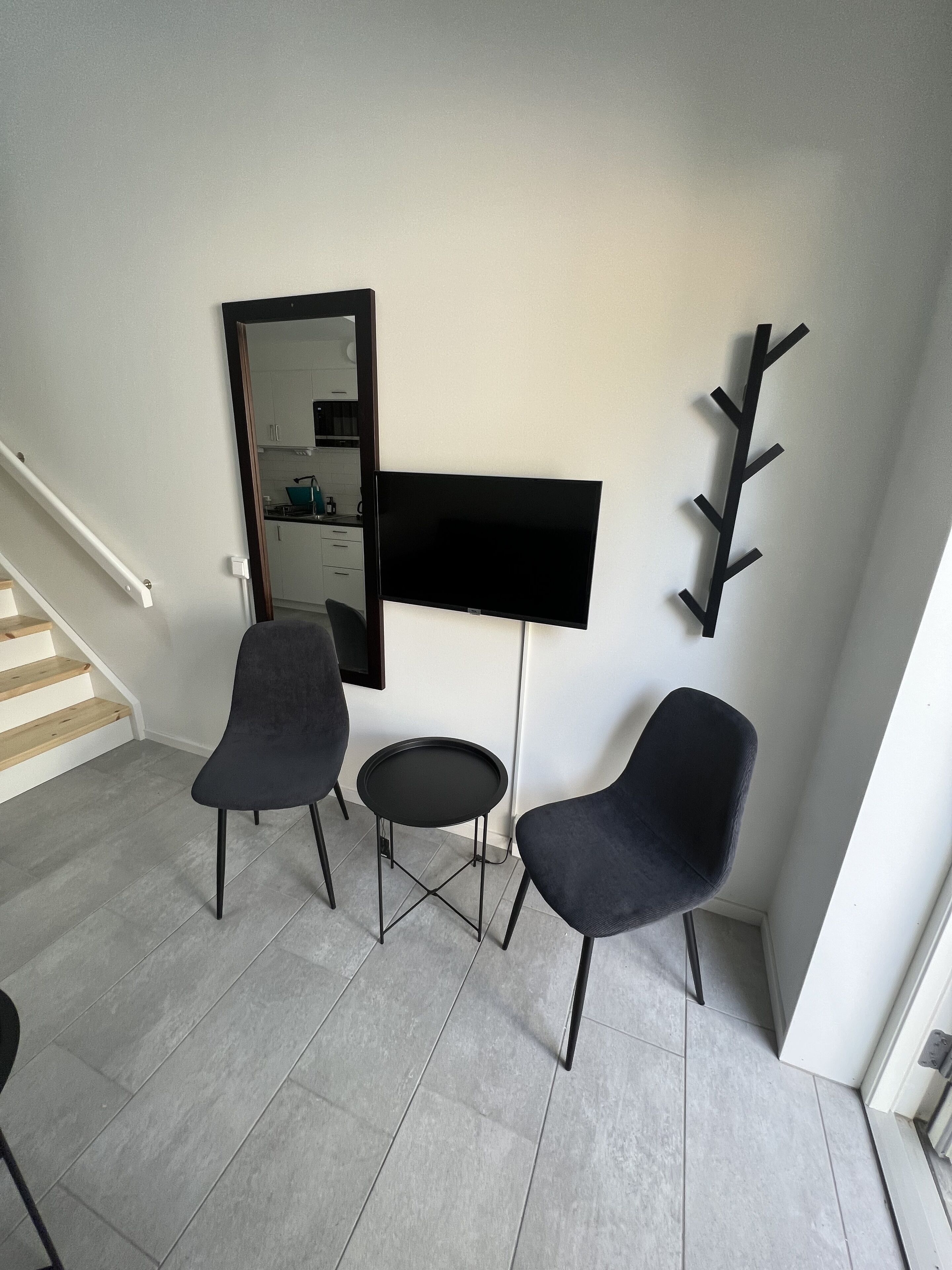 Uma sala de estar moderna com uma televisão de tela plana, duas poltronas escuras, uma mesinha redonda e um porta-roupas.