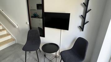 Uma sala de estar moderna com uma televisão de tela plana, duas poltronas escuras, uma mesinha redonda e um porta-roupas.