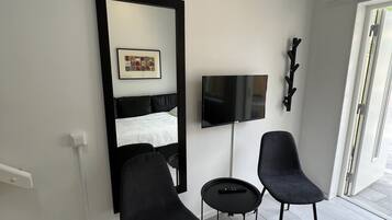 A modern hotel room with a large mirror, a bed with a white duvet, a flat-screen TV, two black chairs, and a small round table.