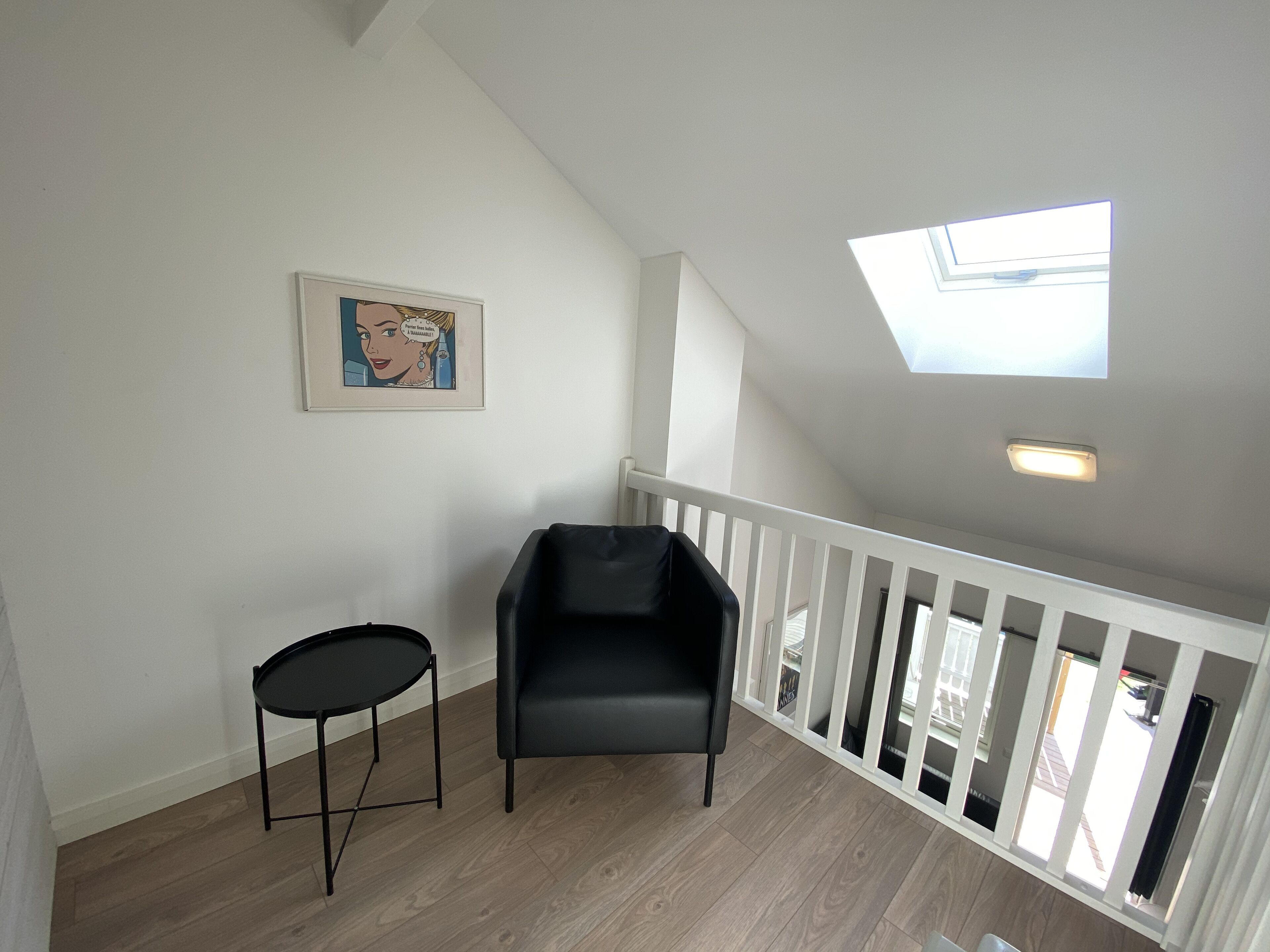 A room with a sloped ceiling, a skylight, a black chair, a small round table, and a framed picture on the wall.