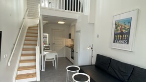 Un interior moderno con una escalera que sube a un altillo, un sofá de cuero negro, dos mesas redondas blancas y una zona de cocina.