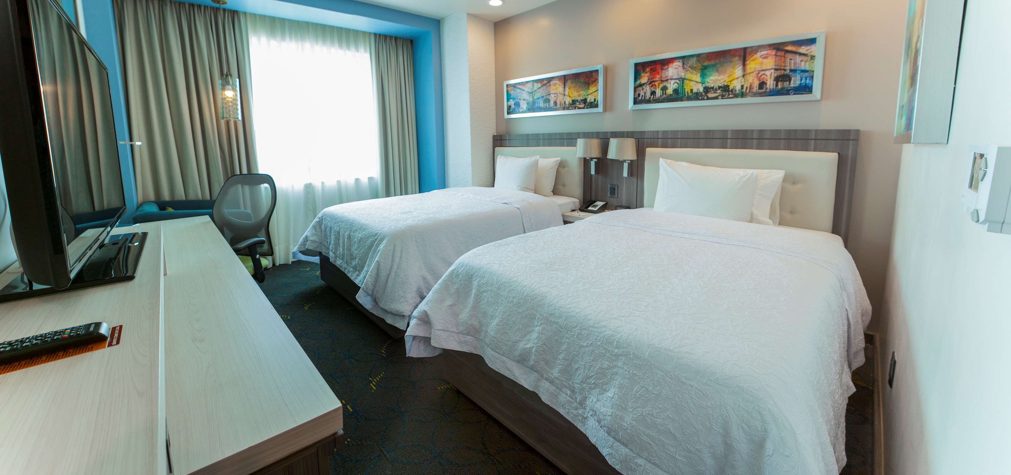 A hotel room with two beds, a desk, a chair, and a TV.