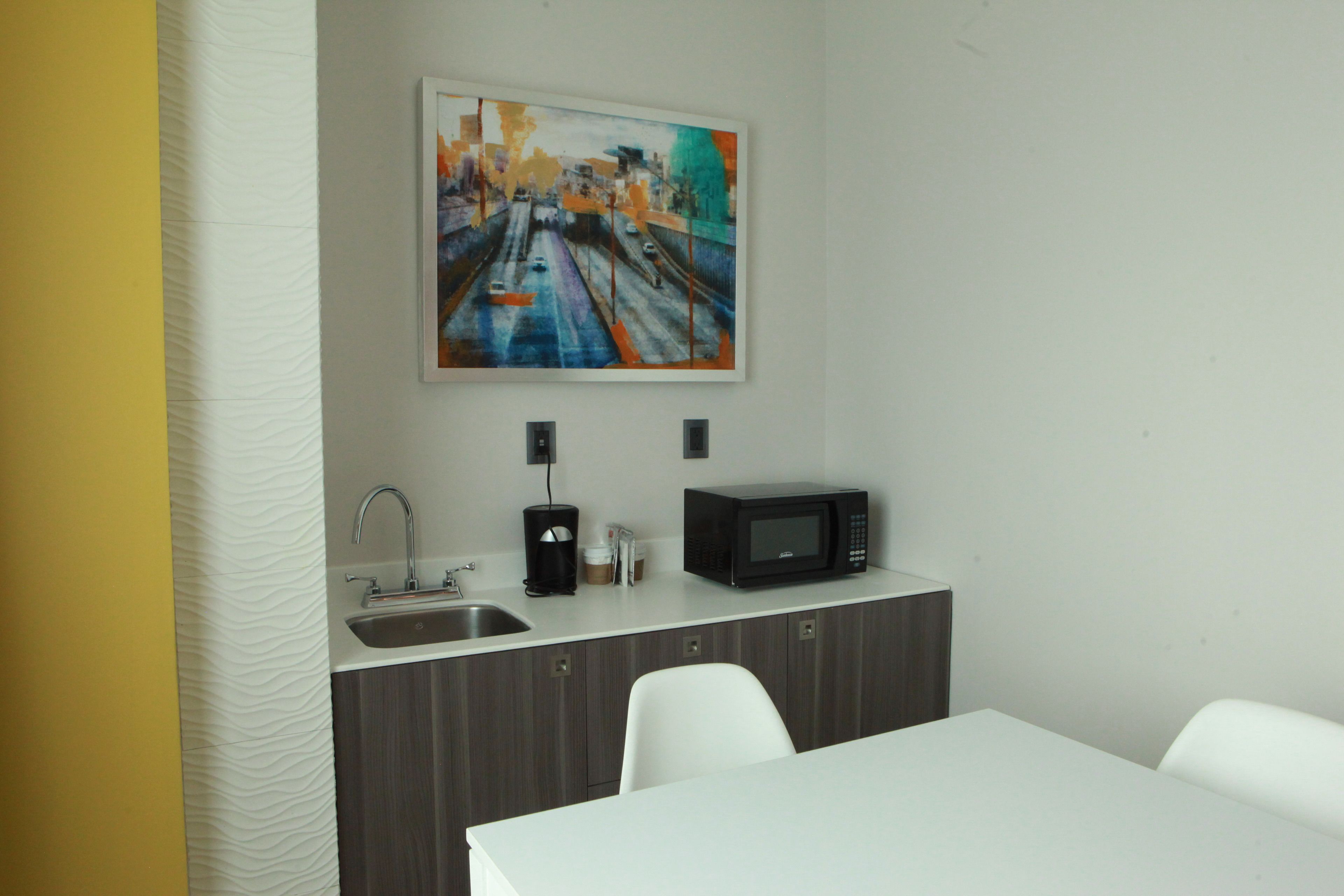 A small kitchenette with a microwave, sink, and coffee maker. There is a painting above the sink and a white table with chairs.