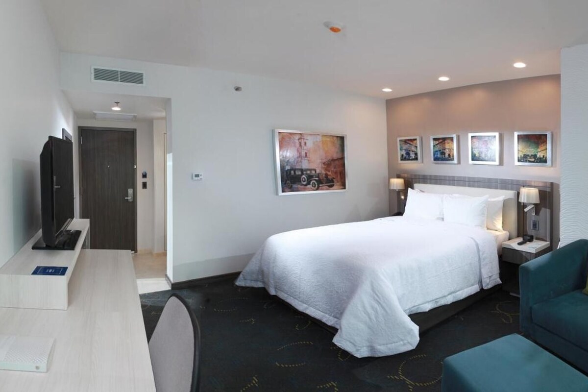 A hotel room with a bed, a desk, a chair, a TV, and artwork on the walls.