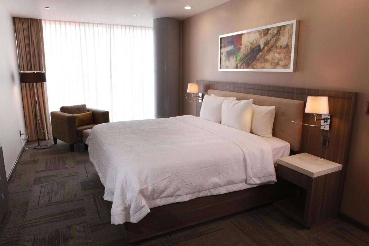 A hotel room with a large bed, a bedside table, a chair, and a painting on the wall.