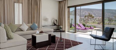 A modern living room with a sofa, coffee table, and a chair. There is a balcony with a view of the mountains.