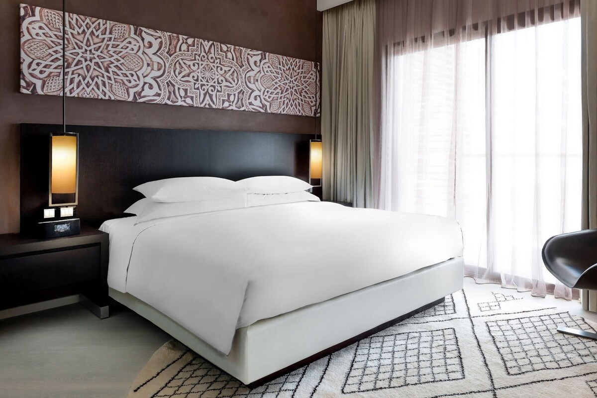 A modern hotel room with a large bed, a nightstand with a lamp, a chair, and a patterned wall decoration.