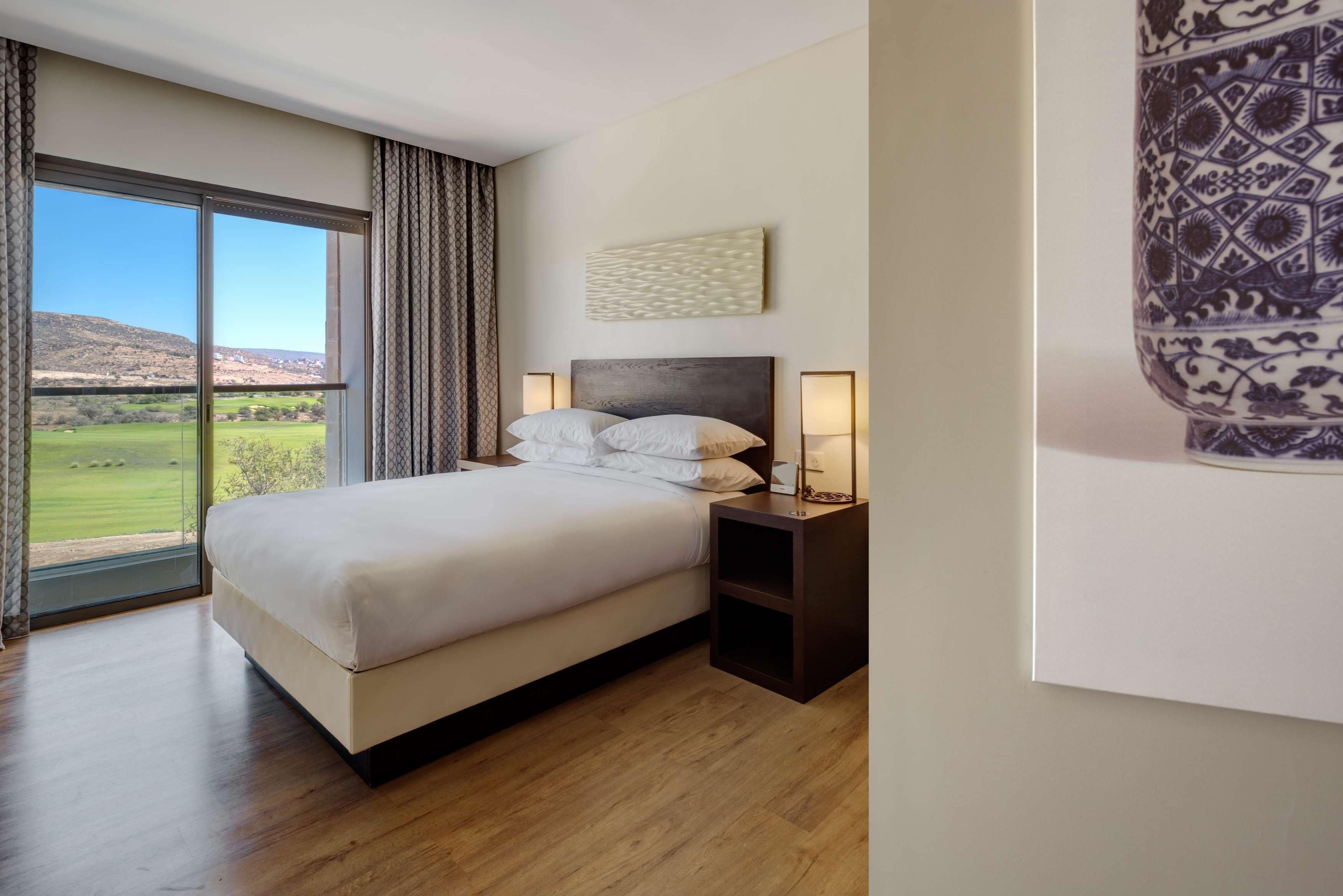 A bedroom with a large bed, a nightstand with a lamp, a view of a green landscape through a large window, and a wall-mounted artwork.