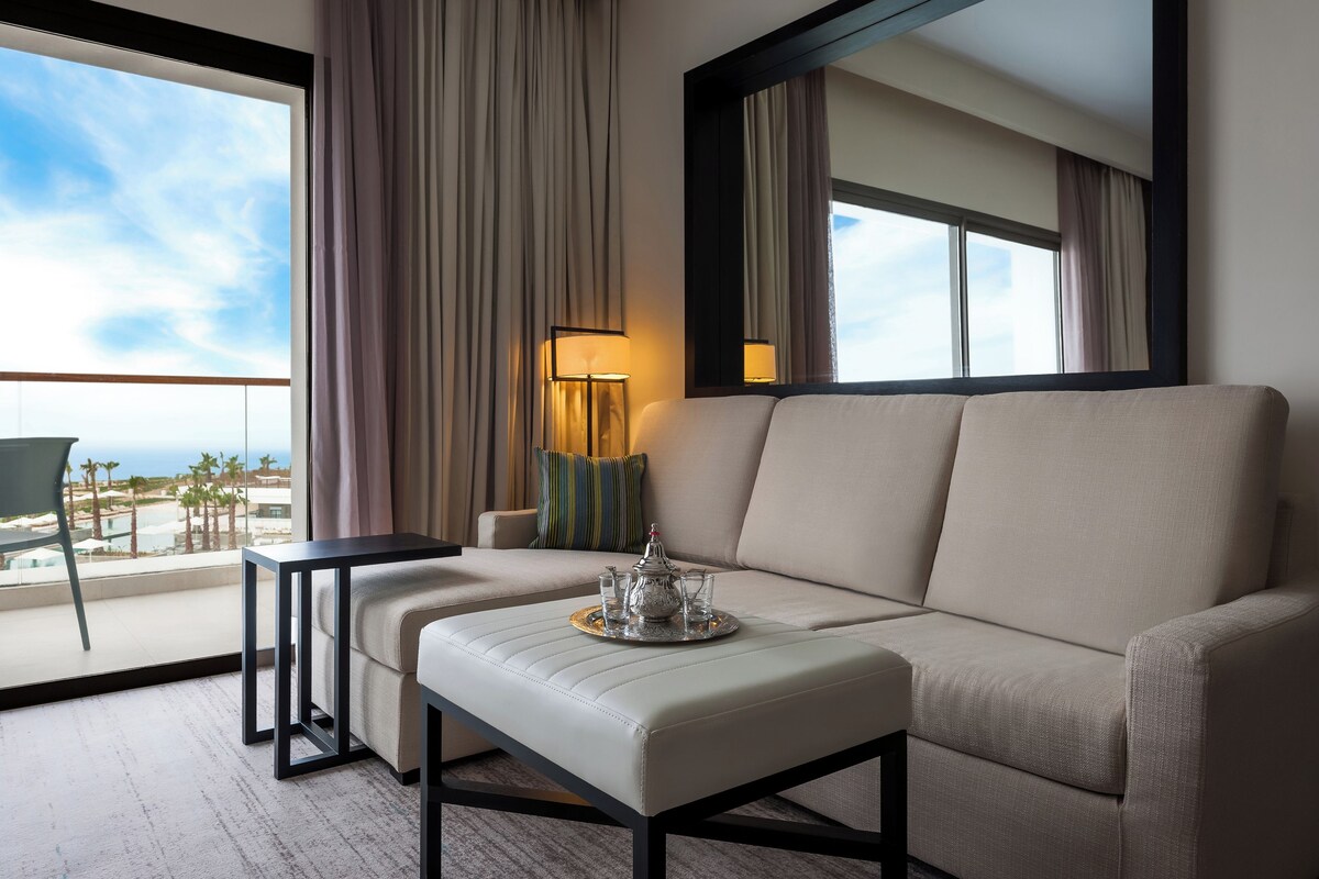 A modern hotel room with a balcony, a sofa, a small table, and a lamp.