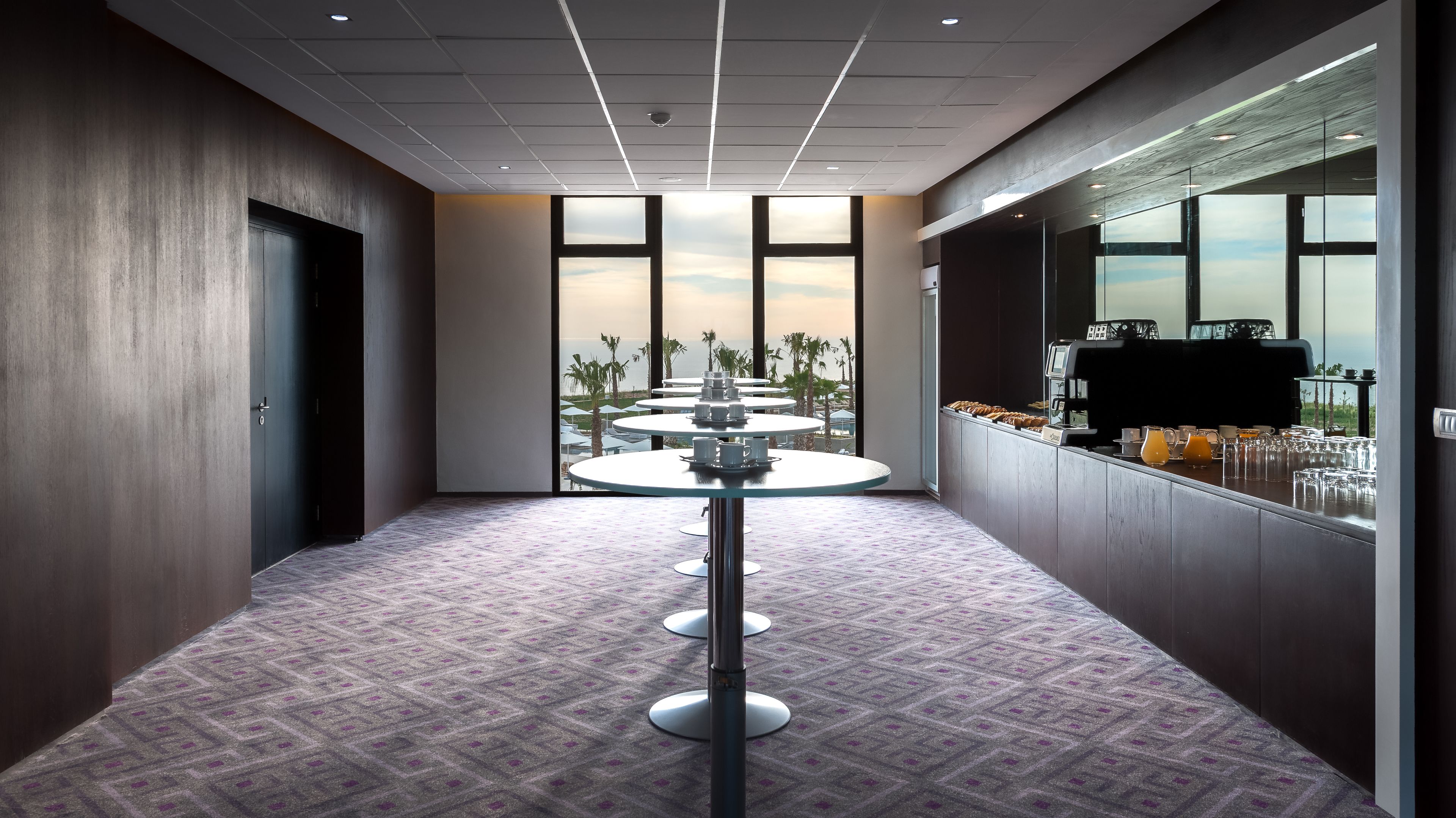 A modern hotel corridor with a patterned carpet, glass tables with trays, and a view of palm trees and the ocean through large windows.