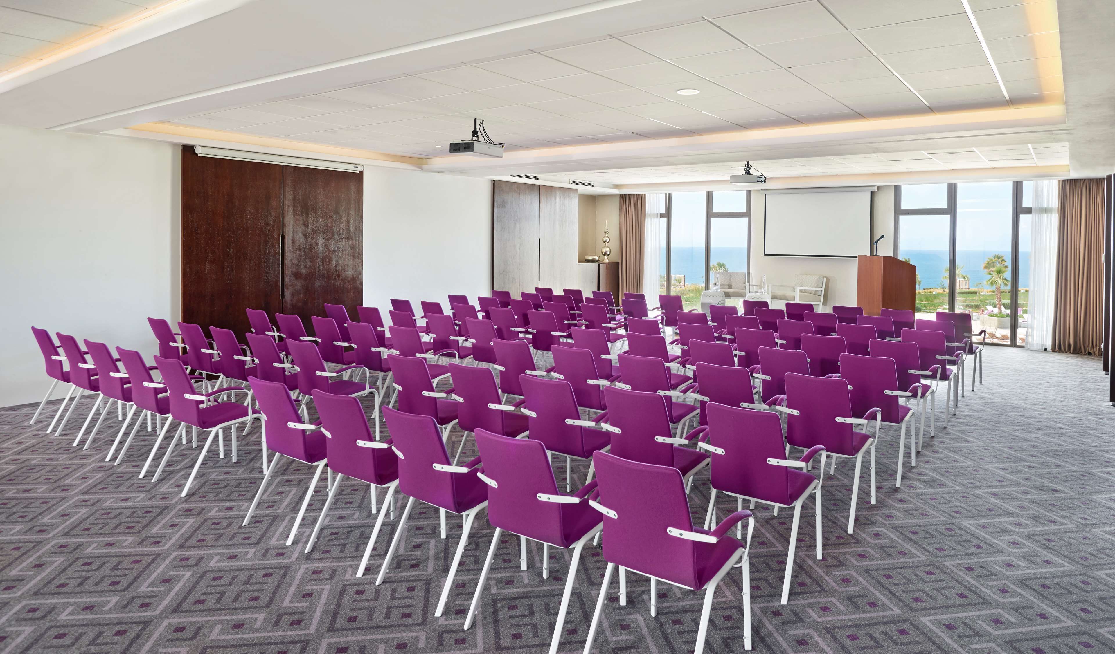 Une salle de conférence avec des rangées de chaises violettes, de grandes fenêtres offrant une vue dégagée et un pupitre.