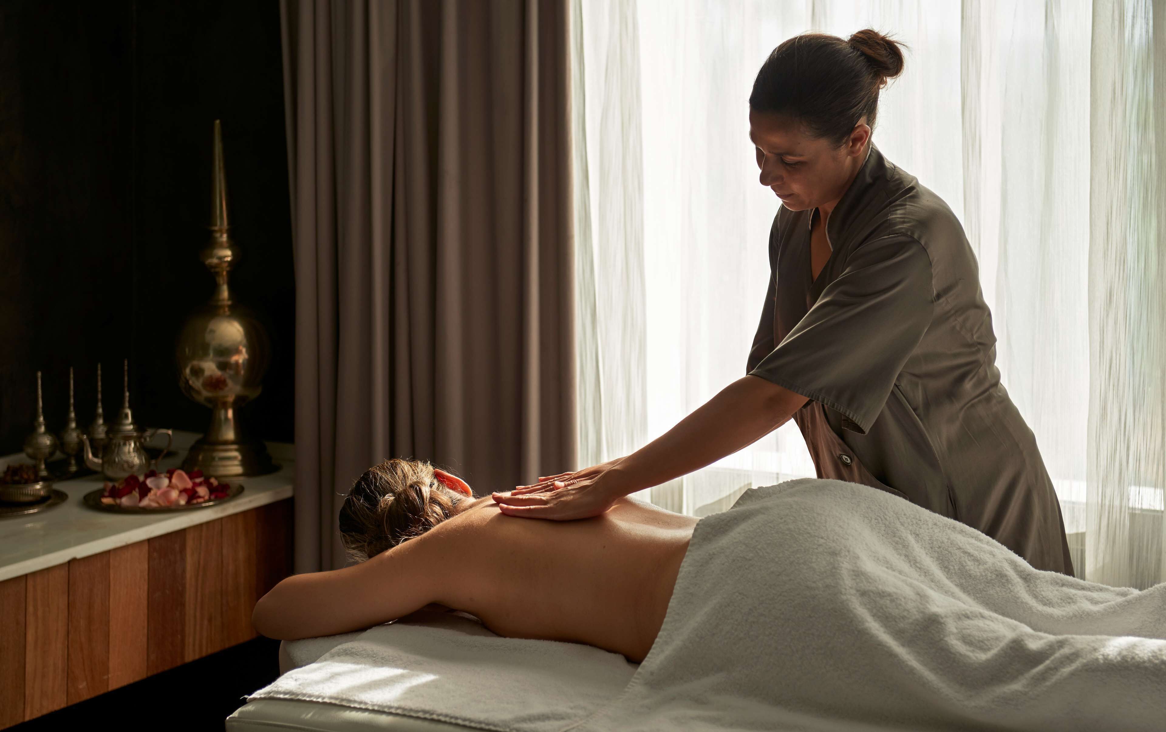 Eine Massageanwendung in einem Raum mit Massageliege, einer Zierlampe und Vorhängen.