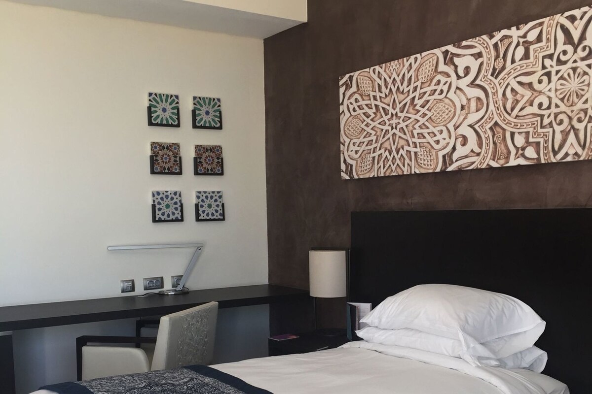 A hotel room with two beds, a desk, a chair, and a wall with decorative tiles.