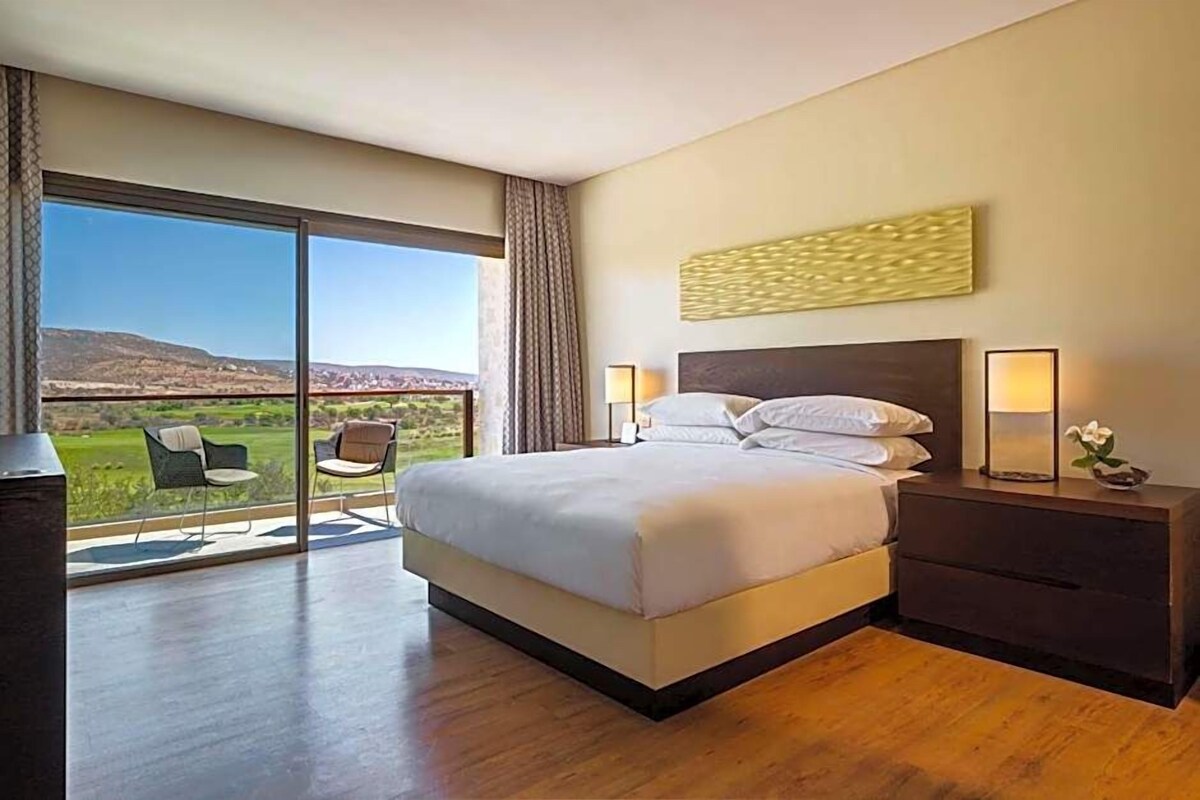 A modern hotel room with a large bed, a nightstand, a lamp, and a view of a green landscape through a sliding door.