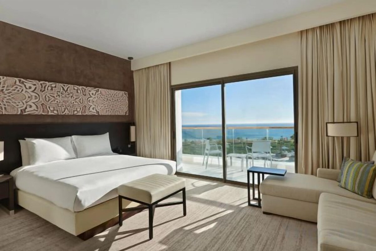 A hotel room with a large bed, a sofa, a balcony with a view, and a nightstand.