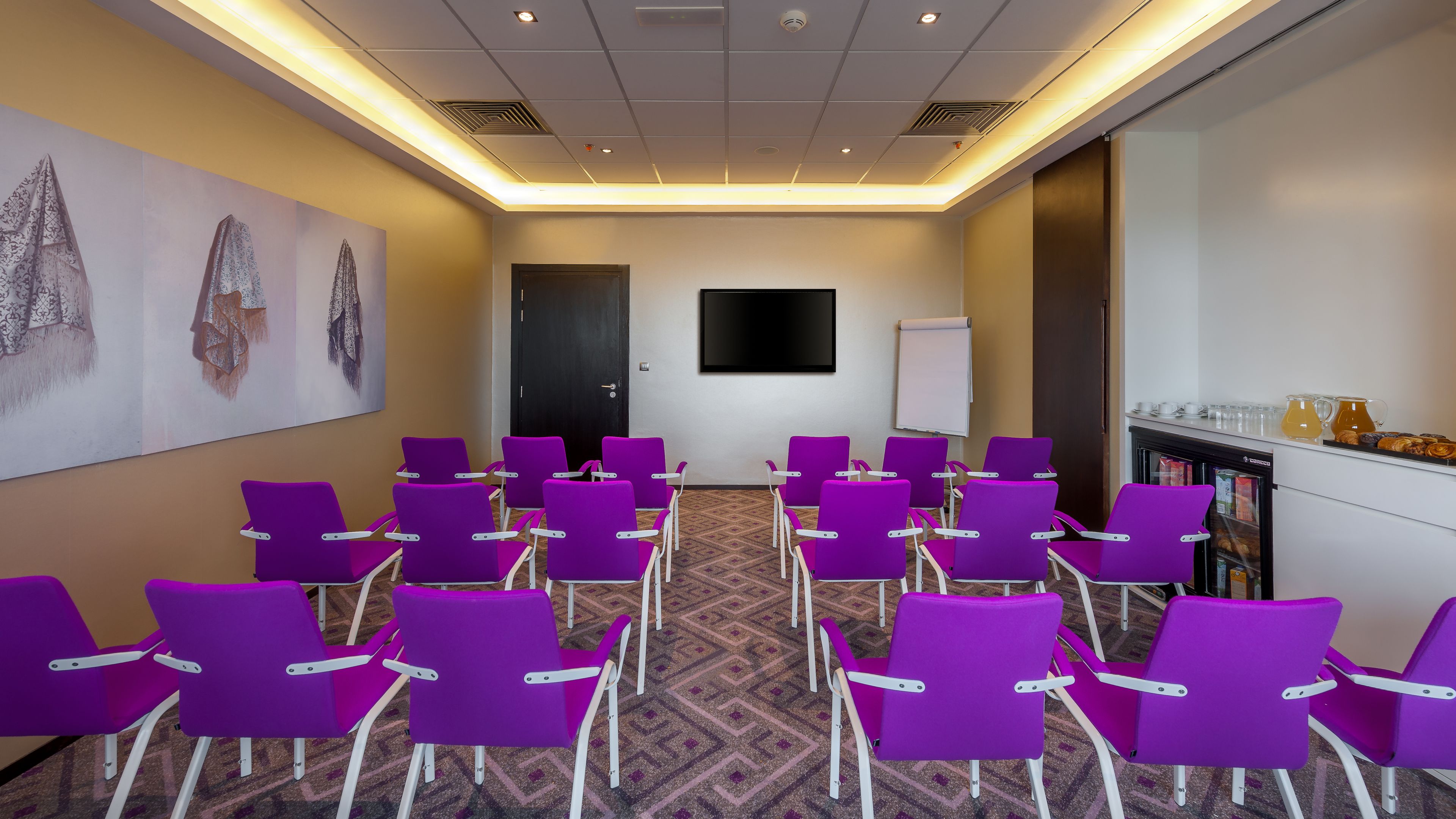 Une salle de conférence avec des chaises violettes disposées en rangées, un tableau blanc et un coin café équipé d’un réfrigérateur.