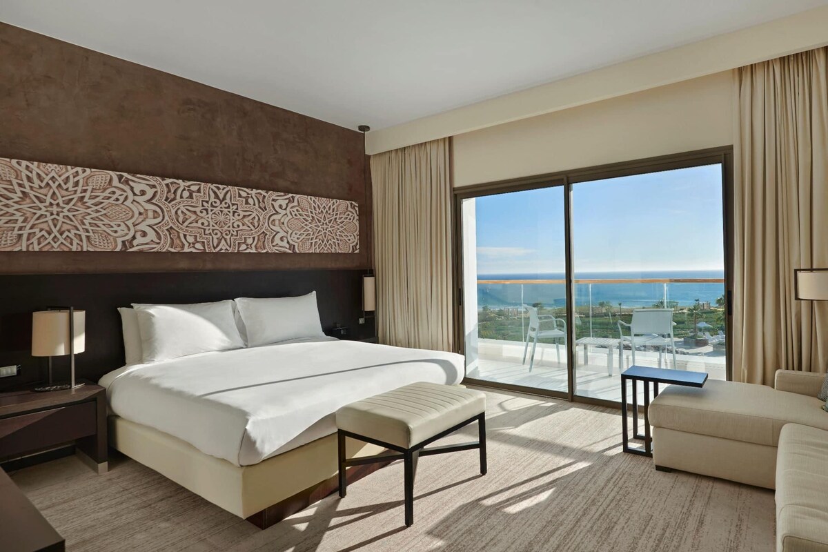 A hotel room with a large bed, a sofa, a balcony with a view of the sea, and a decorative wall panel.